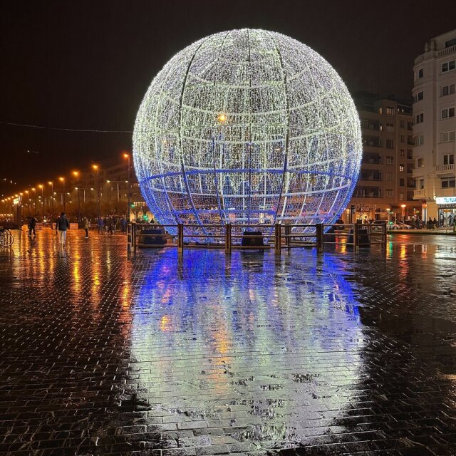 Una esfera enorme tejida con miles de bombillas blancas y azules, levantada en mitad de una plaza nocturna; el suelo mojado le devuelve un planeta paralelo, alargado en tiras de color. Un edificio elegante a la derecha, farolas en hilera a la izquierda, dos paseantes mínimos detrás. La ciudad montó por unos días un astro provisional y la lluvia se encarga de duplicarlo.