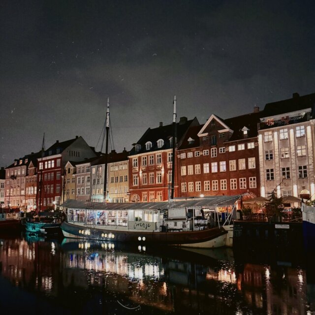 Canal portuario nocturno con casas estrechas de colores y tejados a dos aguas alineadas en el muelle. Dos barcos amarrados en primer plano, uno con el rótulo Oranje, lanzan reflejos rojos y dorados sobre el agua negra. Algunos puertos sólo se entienden cuando se quedan vacíos.
