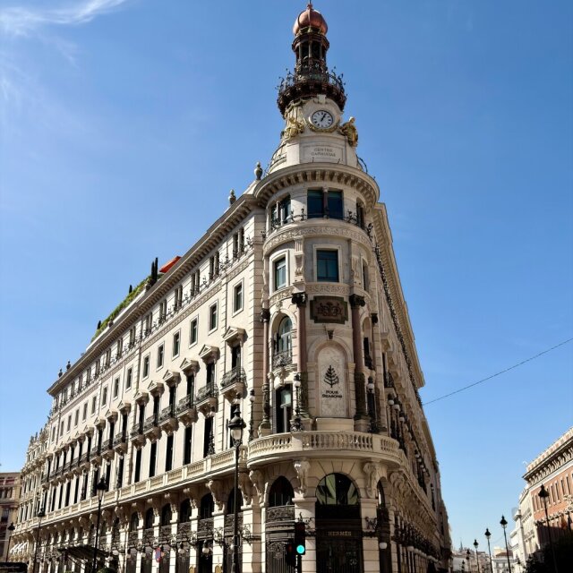 Edificio en esquina aguda, blanco, monumental, coronado por una torrecita con reloj y un capitel de cobre. Pisos de balcones decorados con herrerías negras y, abajo, escaparates de Hermès. Cielo azul plano. Las ciudades guardan sus mejores edificios para los cruces a los que no había una manera fácil de llegar.
