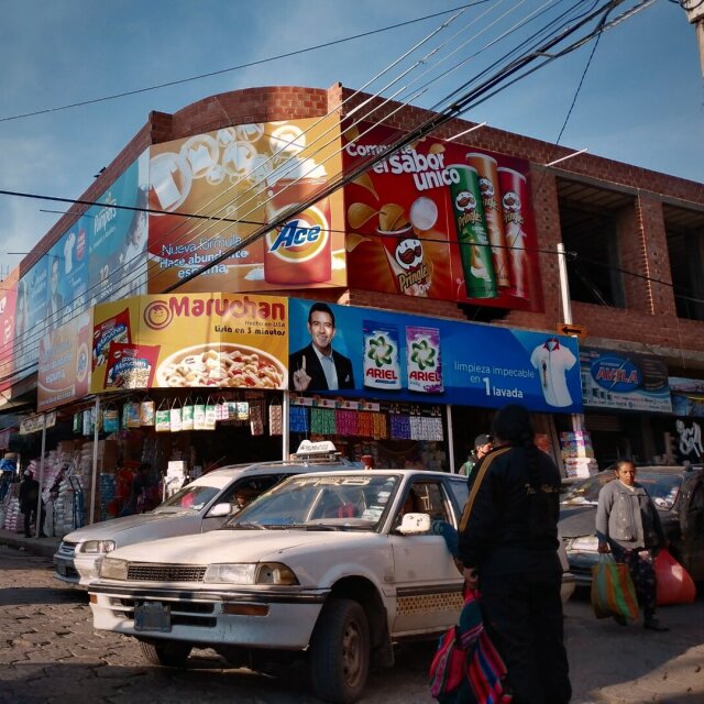 Esquina de una calle con la fachada de ladrillo empapelada de carteles gigantes: Pringles, Ace, Ariel, Maruchan. Un coche blanco viejo aparcado en la cuneta, dos mujeres caminando con bolsas de mercado a rayas. Las marcas globales colonizando, una a una, las paredes de cualquier ciudad. La calle como página de revista a tamaño real.