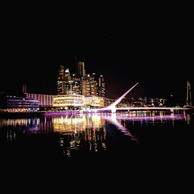 Skyline nocturno al otro lado de un río oscuro: torres de oficinas encendidas, una pasarela de mástil inclinado y tirantes blancos cruzando hacia ellas. El agua, casi negra, devuelve toda la ciudad invertida en hilos verticales de luz. La orilla se abstiene; deja que el reflejo haga el discurso. A veces una ciudad se entiende mejor por su réplica que por su original.