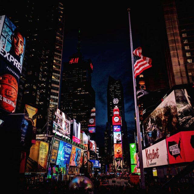 Plaza nocturna iluminada por pantallas gigantes con anuncios de musicales, marcas y carteles de Broadway, una bandera estadounidense a media asta en el centro. Los rascacielos quedan reducidos a estructura entre el ruido visual. Algunas plazas son ya pantalla pura.