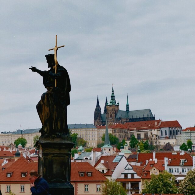 Estatua de un santo con cruz dorada en alto y dedo apuntando, recortada contra una vista panorámica de tejados rojos y una catedral gótica con cúpulas verdes al fondo. Un visitante asoma por la base sin levantar la vista. Las indicaciones más antiguas siguen señalando lo mismo.