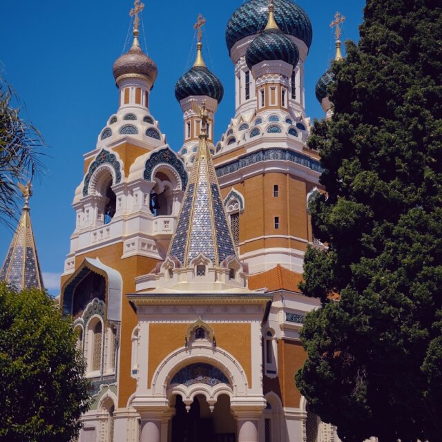 Catedral con cúpulas en bulbo de teja vidriada azul y dorada que asoman sobre fachadas de ladrillo naranja y mármol blanco. Una palmera y un ciprés enmarcan el conjunto contra un cielo abierto, sin nubes; en el atrio, una figura solitaria sube los escalones. Una arquitectura del frío, varada bajo un sol del sur.