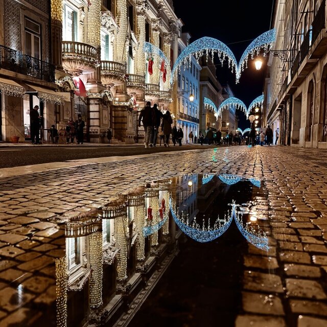 Calle empedrada por la noche, mojada de lluvia. A la izquierda, una fachada barroca cubierta entera de bombillas amarillas como si la hubieran vestido para una ópera; sobre la calzada, arcos azules de luces colgados de un edificio a otro. Y abajo, en el agua de los adoquines, todo doblado al revés con una nitidez incómoda. La lluvia hace doble exposición sin pedir permiso.