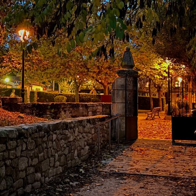 Entrada de un parque nocturno con farolas de gas encendidas entre castaños amarillos. Un portón de hierro con pilar de piedra rematado en bola separa la calle del paseo cubierto de hojas. El parque deja ver lo suyo cuando ya nadie pasa.