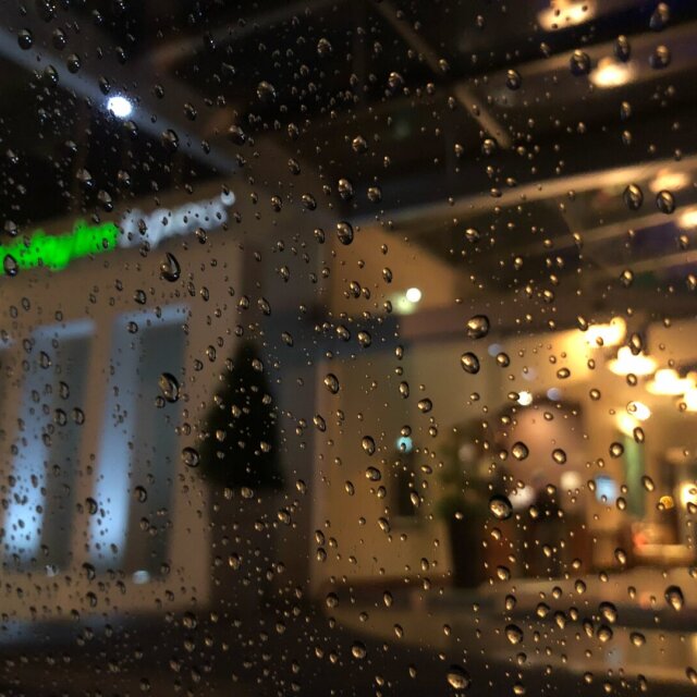 Cristal mojado en primer plano: cientos de gotas perfectamente esféricas, cada una funcionando como lente diminuta, distorsionan a su modo el fondo desenfocado. Detrás se adivinan ventanas iluminadas en cálido, un árbol de Navidad pequeño y un rótulo verde. Nada está nítido, nada importa. La frontera del enfoque es también la frontera del recuerdo.