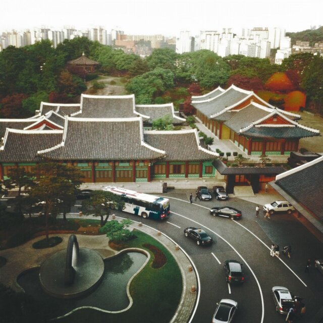 Vista aérea de un palacio coreano: tejados de pagoda en cascada, vigas lacadas en rojo y verde, patios despejados. En el horizonte, un mar de rascacielos pálidos. Entre uno y otro, un autobús turístico estacionado y dos sedanes negros esperando. La dinastía sobrevive como isla rodeada de torres genéricas. Lo que era el centro acabó siendo plaza interior.