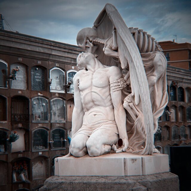 La escultura del beso de la muerte: un esqueleto alado se inclina y besa en la frente al joven semidesnudo, arrodillado, que echa la cabeza atrás como quien recibe un alivio. La mano huesuda ya posa sobre la cadera; la otra ala, abierta, empuja al cielo. Detrás, las celdillas del cementerio repiten la misma gramática: nicho, fecha, nombre. La rendición no es derrota, es sólo cuestión de postura.