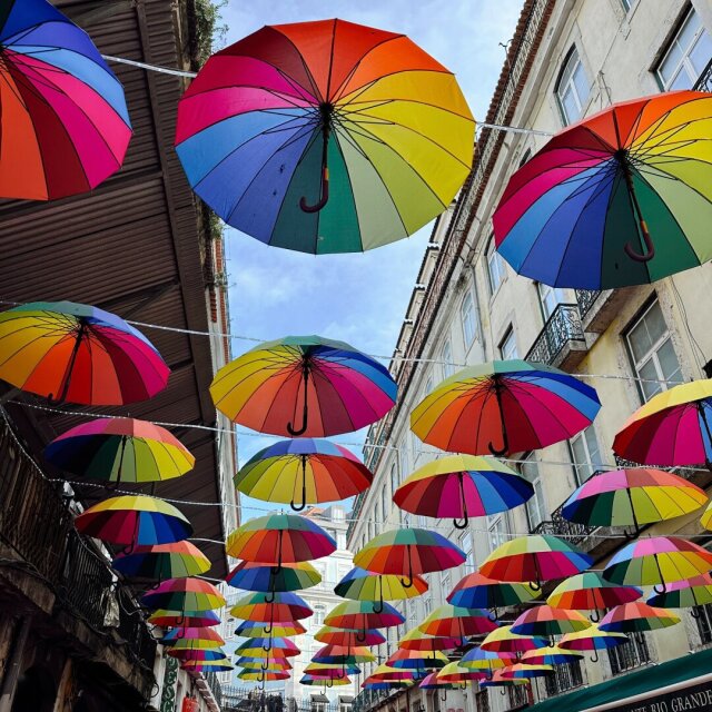 Una calle entera techada con cientos de paraguas multicolores colgados de cuerdas: arcoíris a granel sobre la cabeza, fachadas a derecha e izquierda con balcones de hierro y un restaurante al fondo. Los transeúntes apenas se ven, eclipsados por el dosel de tela. El resguardo convertido en instalación: la lluvia, si llega, va a tener que pedir cita.