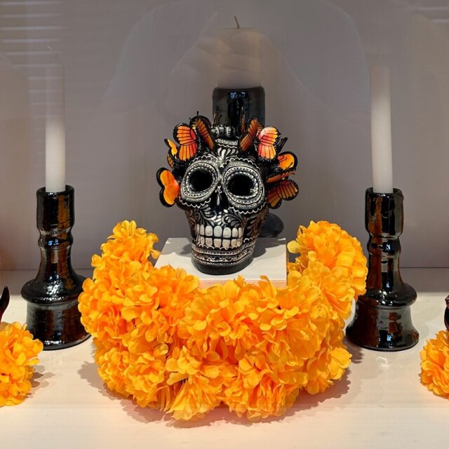 Tres calaveras de cerámica esmaltada en una vitrina: la del centro lleva mariposas monarca anidadas en la cabeza; las laterales, llamas pintadas y guirnaldas. Entre ellas, una alfombra densa de cempasúchil naranja y dos velones negros encendidos. Todo limpio, nada solemne. La muerte aquí no asusta: se peina, se viste de naranja y se sienta a esperar a los suyos como quien recibe en casa.