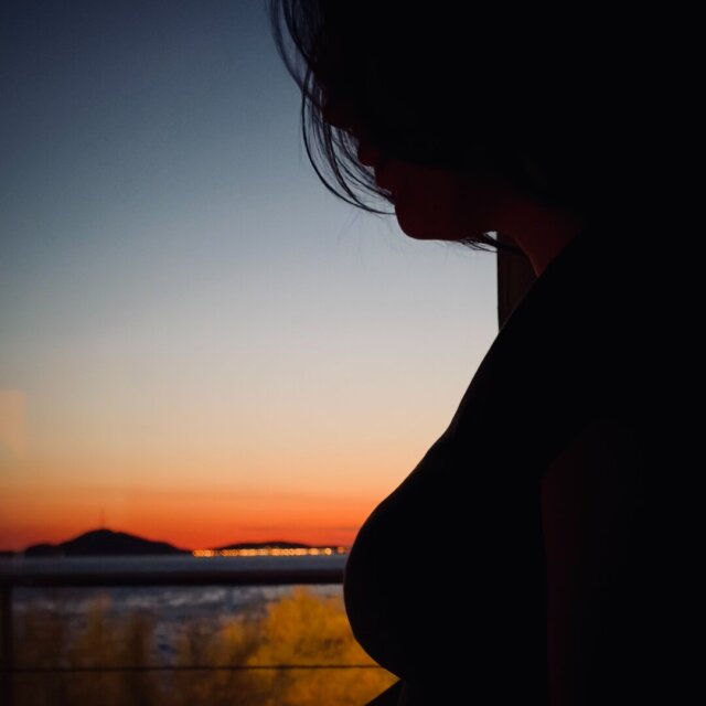 Silueta femenina recortada contra un ventanal: pelo, perfil bajado, hombro y curva del torso en sombra plena. Detrás, el horizonte se va degradando del naranja brasa al azul de noche, con una isla mínima y un puñado de luces lejanas haciendo collar sobre el agua. La oscuridad de dentro es más densa que la de fuera. Hay momentos en los que el cuerpo se vuelve frontera, no figura.
