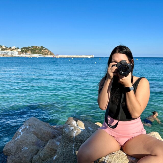 Mujer en bañador rosa sentada sobre una roca al borde del mar, descalza, levantando una cámara Sony para fotografiar al fotógrafo. Pelo al viento, agua color esmeralda detrás, una cabeza nadando a la derecha y una costa baja con caserío blanco al fondo. El visor le devuelve a quien la mira la mira de vuelta. Cuando la mirada se duplica, deja de haber sujeto y objeto.
