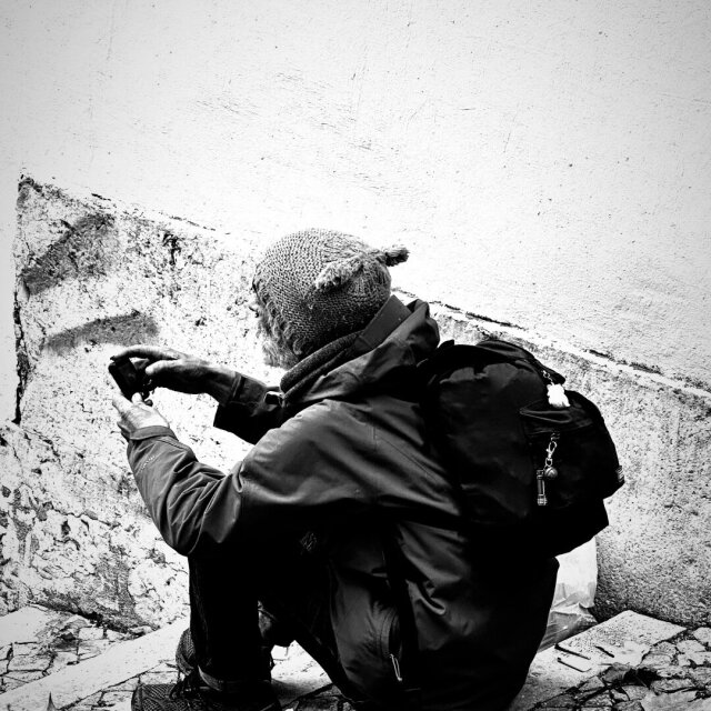 Hombre acuclillado de espaldas contra una pared blanca y descascarillada: gorro de lana con borlas, abrigo grueso oscuro, mochila con un llaverito colgando. Sostiene una pequeña cámara con las dos manos sucias y se inclina a apuntar hacia algo que no vemos. Calle empedrada con hojas secas. Incluso quien parece no tener nada elige todavía a qué prestar atención.