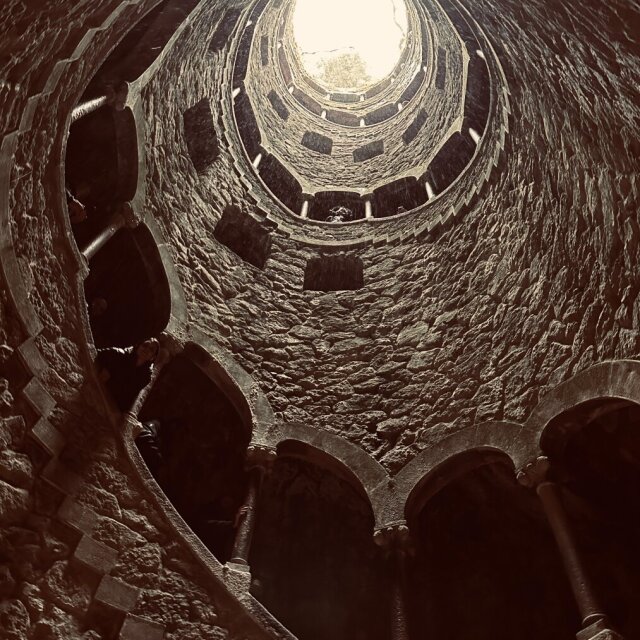 Pozo iniciático visto desde abajo: una espiral descendente de piedra labrada que sube en torno a un eje vacío hasta abrirse en un óculo de luz, con un árbol asomado al borde como advirtiendo que arriba aún hay mundo. En las galerías laterales se intuye gente diminuta. Bajar para ascender es solo gramática del mismo gesto.