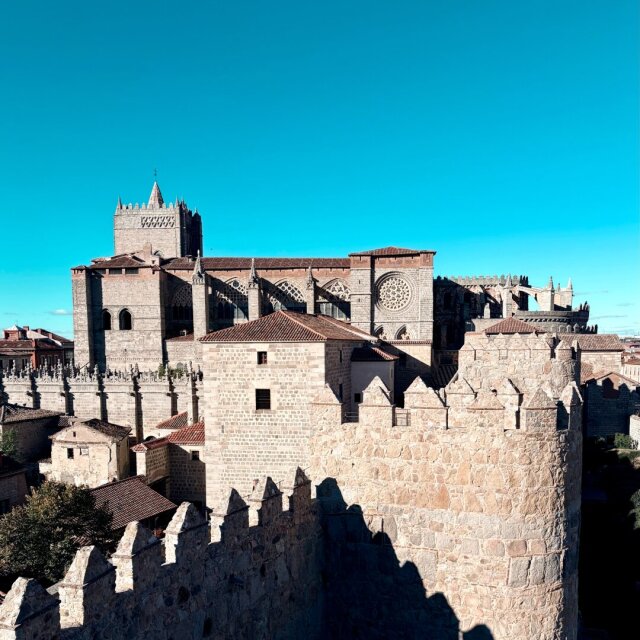 Almena de muralla medieval en primer plano sobre la que asoma una catedral gótica con rosetón y cimborrio fortificado. El cielo azul lavado deja la piedra entera para sí misma. Las ciudades amuralladas se acostumbraron pronto a caber en su propio perímetro.