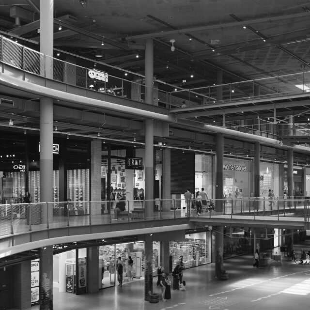 Interior de un centro comercial en blanco y negro: estructura metálica vista, barandillas con malla, vigas y conductos al aire. Dos plantas de tiendas se asoman al hueco central; abajo, las personas, apenas reconocibles, se desparraman por el suelo brillante. La luz cenital es la de un sitio donde nadie sabe qué hora es.