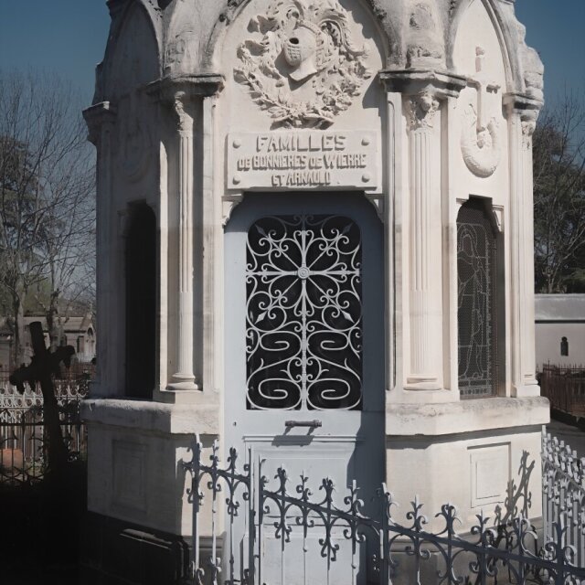 Mausoleo neoclásico en mármol blanco con escudo heráldico tallado en el frontón y reja de hierro forjado en arabescos sobre la puerta. Lo flanquean otras tumbas y una cruz negra de hierro asoma a la izquierda. La filigrana del cierre dura más que el apellido grabado encima.