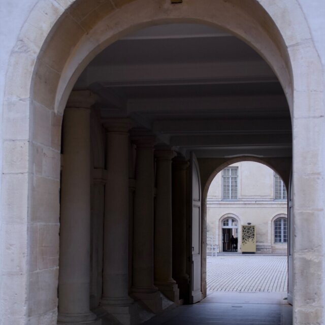 Vista enfilada de dos arcos: uno grande en primer plano enmarca el segundo, más pequeño y al fondo, que se abre a un patio empedrado. Entre ambos, pilastras estriadas y un suelo enlosado componen una geometría limpia. Cada arco anuncia el siguiente; ningún umbral termina del todo.