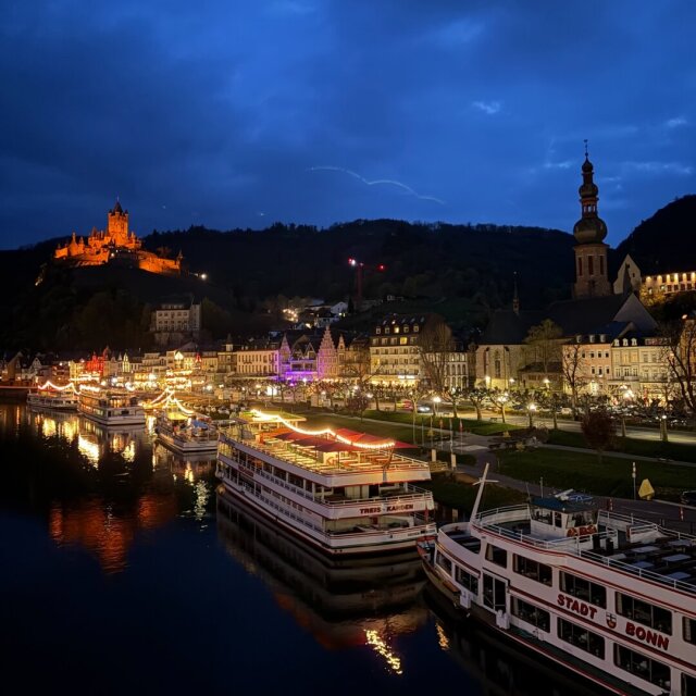 Pueblo a orillas de un río al anochecer: en lo alto, un castillo iluminado de naranja preside la silueta del valle; abajo, dos barcos de pasaje amarrados se reflejan en el agua negra. Entre el castillo y el muelle, casas con luces interiores y una iglesia de campanario bulboso ordenan la línea del paseo. La hora azul aguanta mientras los barcos esperan a que alguien suba.