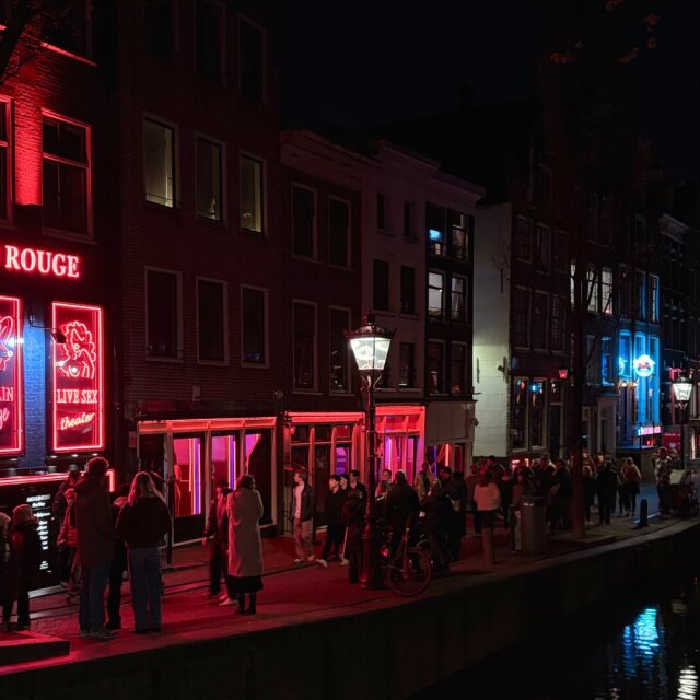 Una calle nocturna teñida de rojo: rótulos de neón anuncian un cabaret y una sex shop sobre fachadas de ladrillo, y un grupo de personas se detiene a mirar las puertas iluminadas. A la derecha, un canal devuelve los reflejos rojos y azules en cintas alargadas. La iluminación es la del distrito que vive del exceso ajeno.