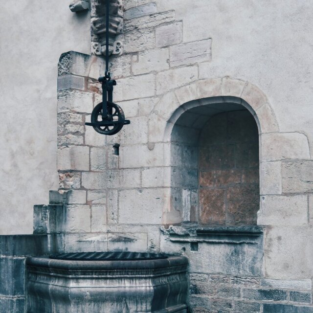 Fuente mural antigua: una cabeza de león tallada en piedra alimenta un pitorro de hierro forjado con válvula y rueda, y vierte sobre una pila circular gastada. A la derecha, una hornacina vacía abre un hueco oscuro en la pared encalada. La fuente sigue funcionando aunque ya no haga falta.