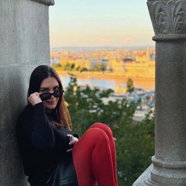 Una mujer joven, sentada entre dos columnas de piedra clara, mira hacia la cámara mientras se baja las gafas de sol. Suéter negro, falda corta de cuero, medias rojas y zapatos a juego: la nota de color queda concentrada en sus piernas. Detrás, una ciudad ribereña se enciende en luz de tarde mientras ella decide cuándo mirar.