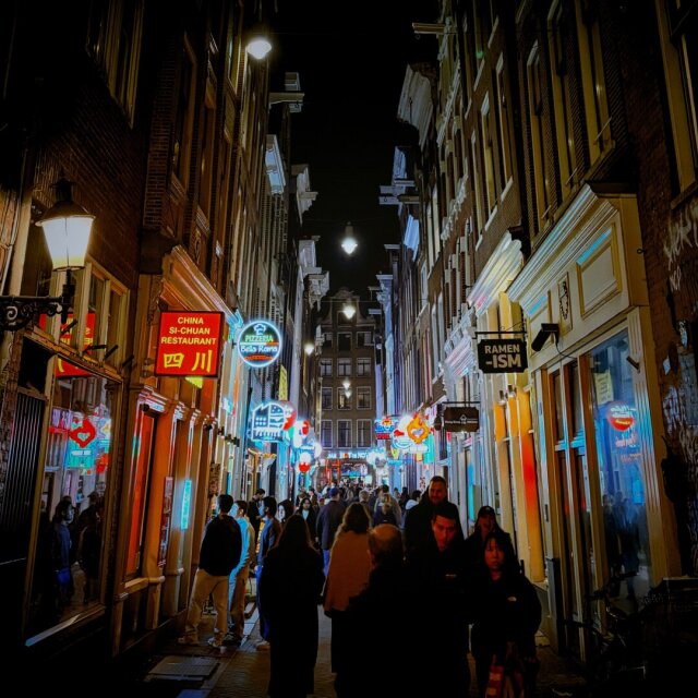 Callejón estrecho de noche con neones de restaurantes asiáticos a ambos lados: rótulos rojos, azules y blancos compiten por la mirada del paseante. Una multitud avanza a contraluz, las caras hundidas en sus móviles o en los letreros. La calle huele a fritura y a sirope dulce sin necesidad de probar nada.
