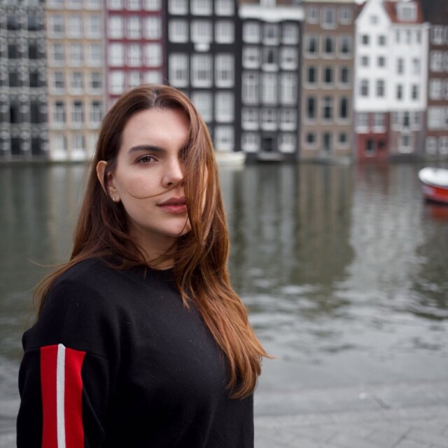Retrato de una mujer joven; un mechón cruza su cara y los ojos se sostienen en cámara sin sonrisa de cortesía. El suéter negro tiene una franja roja vertical en el hombro. Detrás, fachadas estrechas y altas se alinean en hilera sobre un canal de aguas quietas, con un bote rojo asomando al margen.