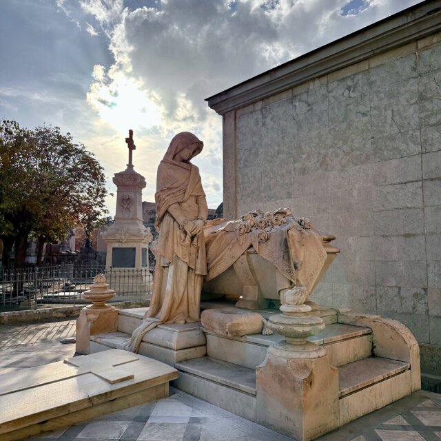 Conjunto funerario en un cementerio: una figura femenina velada, tallada en piedra ocre, se inclina junto a un sarcófago rematado con un ramo de flores en relieve. Detrás se eleva otro panteón coronado por una cruz, y entre las nubes el sol abre un haz directo. La piedra parece más viva que las flores que ya no están.