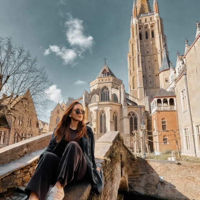 Una mujer joven sentada en un parapeto de piedra al borde de un canal estrecho; lleva gafas de sol y la cabeza vuelta hacia un lado, en un gesto pausado. Detrás, una iglesia gótica de ladrillo levanta una torre afilada hasta lo alto del cuadro, con casas de tejado escalonado a la izquierda. Una postal con alguien dentro que no posa para ella.