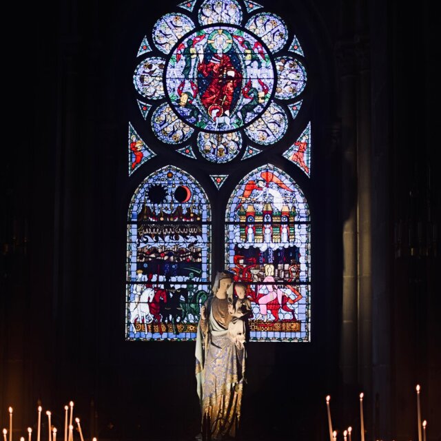 Interior gótico en penumbra: una vidriera con rosetón y dos lancetas figurativas se enciende en azules y rojos contra la pared oscura. Bajo ella, una estatua de Virgen con Niño se sostiene en silueta sobre un mar de velas votivas que arden a sus pies. La luz de cera dura menos que la luz pintada de hace siglos.