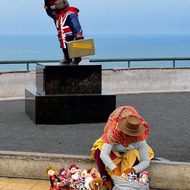 Una estatua de Paddington con la bandera del Reino Unido en el pecho preside un paseo marítimo desde su pedestal de piedra; detrás, el océano gris se pierde sin barcos a la vista. A los pies del monumento, una mujer con sombrero ancho de fieltro y pollera de varios colores organiza muñecos de tela en una cesta para venderlos. Dos turismos del mismo paisaje, sin tocarse.
