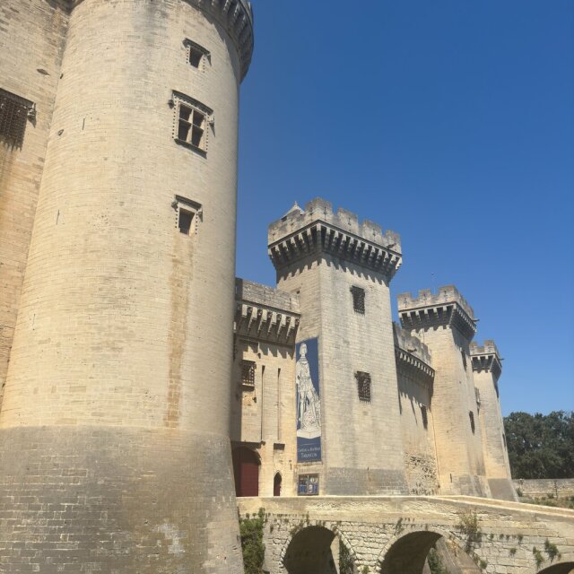 Castillo de piedra clara visto desde abajo: una torre redonda enorme a la izquierda y, detrás, varias torres cuadradas almenadas con saeteras estrechas como ojos pequeños. Un puente de piedra de tres arcos cruza un foso seco hasta la entrada. Cielo azul rotundo. Las fortalezas intactas son siempre una imagen de fracaso útil: las que no llegaron a tener que defenderse.