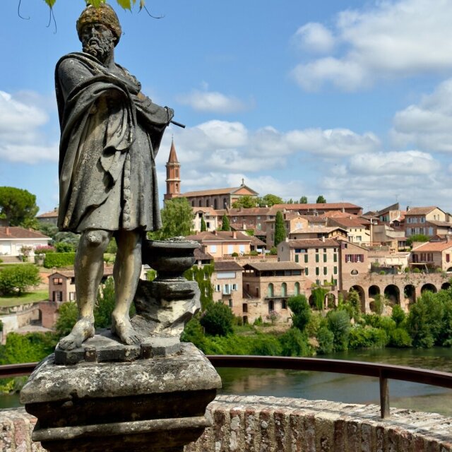 Estatua de bronce ennegrecido sobre un pedestal de piedra: un explorador con turbante, capa larga y bastón, mirando a un horizonte que ya no le incumbe. Detrás, un pueblo de tejados rojos baja hacia el río, con un campanario destacado y un puente de arcos. El monumento envejecido acaba siendo más parte del paisaje que figura sobre él.