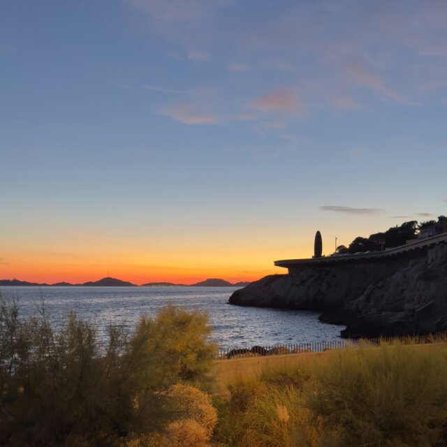 Atardecer en cala estrecha: mar oscuro casi quieto, una pequeña carretera con voladizo blanco sobre la roca, vegetación seca y arbustos en primer plano, dos o tres islas mínimas en el horizonte y un cielo que va del naranja del fuego al azul de la madrugada anterior. El sol se va y el mirador se queda mirando hacia donde ya no hay nada.