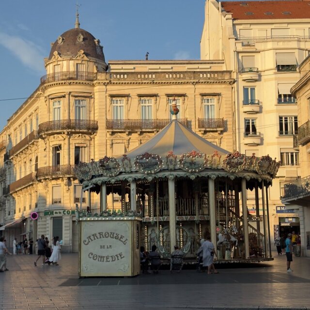 Plaza al atardecer dorado: el Carrousel de la Comédie con su tienda de tela rayada, caballitos pintados y bombillas encendidas; detrás, un edificio haussmaniano con cúpula azul oscuro y balcones de hierro. Una paloma sola en mitad del enlosado. Cuando el carrusel cierra, la plaza vuelve a ser piedra y a recordar lo callada que es de costumbre.