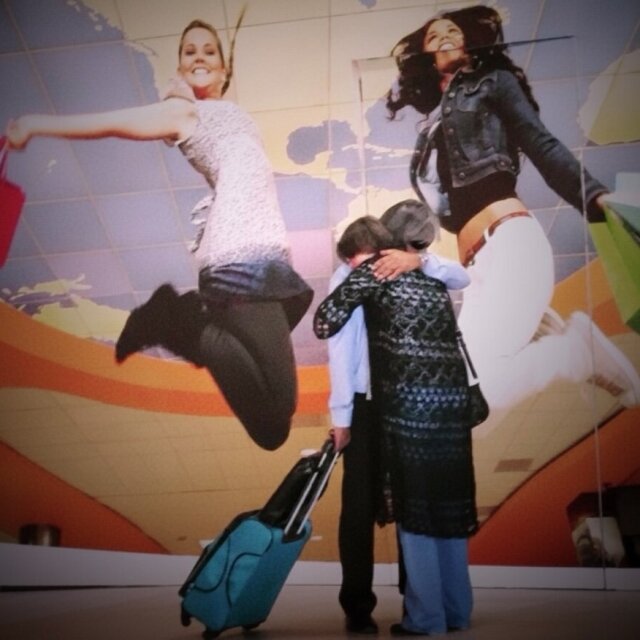 Aeropuerto: en primer plano, dos personas se abrazan junto a una maleta turquesa; detrás, un mural publicitario gigante con dos mujeres saltando con bolsas de compras sobre un mapa del mundo. El gesto verdadero queda más pequeño que el gesto inventado. Las despedidas reales nunca caben en un cartel.