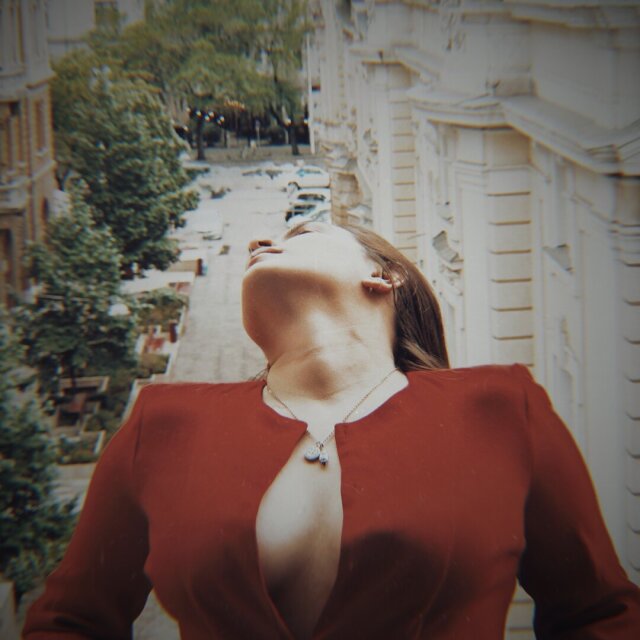 Composición vertical en tonos sepia: una joven con vestido rojo inclina la cabeza hacia atrás, su rostro se funde con una vista en picado de una calle arbolada con coches aparcados y fachadas señoriales. La unión entre cuerpo y ciudad parece costura, no superposición. Algunas calles también se respiran hacia adentro.