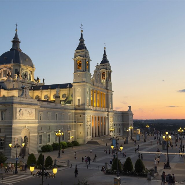 La catedral de la Almudena al atardecer: cúpula azul oscuro con linterna, dos torres barrocas rematadas en agujas y un costado largo con balconadas. La piedra clara recibe el último amarillo del sol. Plaza con farolas ya encendidas y gente paseando minúscula. La catedral nueva imitando vejez para que la traten con el respeto que dan los siglos.