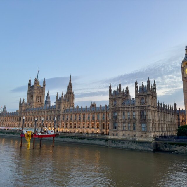 El palacio de Westminster visto desde el río al atardecer: una hilera larga de pináculos góticos color miel, ventanas ojivales y, a la derecha, la torre de Big Ben recibiendo todavía la última luz dorada cuando la fachada ya está en penumbra. El Támesis turbio en primer plano. El imperio sigue iluminado por hábito, no por necesidad.