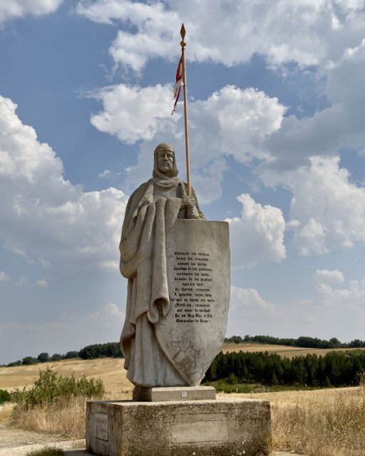 Estatua de piedra clara del Cid Campeador en pleno campo castellano: cota de malla, manto largo, lanza muy alta con pendón rojo y blanco, escudo enorme apoyado en el suelo en cuya superficie hay grabada una estrofa entera del Cantar. Cielo de nubes blancas a granel, trigales segados y un bosquete al fondo. El héroe convertido en escudo de su propio texto.