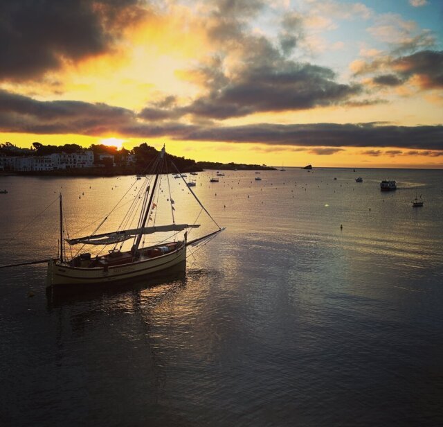 Velero tradicional amarillo amarrado en una bahía al amanecer, mástil sin velas izadas. Otros barcos puntean el agua tranquila hasta una costa lejana de casas blancas. El sol asoma entre nubes, derramando una franja dorada sobre el mar. Hay puertos donde el día empieza despacio porque el oficio sigue siendo esperar.