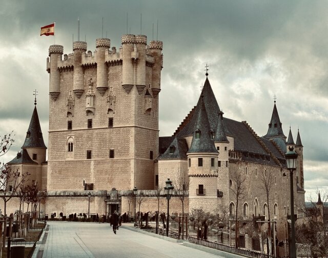 Alcázar fortificado sobre un espolón, torre del homenaje rematada en cubo con torrecillas almenadas y bandera en lo alto, cuerpo principal con tejados cónicos de pizarra a la derecha. Cielo plomizo, fila de árboles esqueléticos en el paseo. Más castillo de cuento que castillo de batalla: la silueta gana siempre a la función.