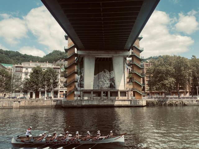 Vista desde abajo de un puente urbano de vano amplio, en el muro central un mural pictórico difícil de leer en escorzo. Por el río pasa una trainera de ocho remeros con patrón al mando, casco blanco con letras rojas. Edificio decimonónico a un lado, ribera urbana al otro. La ciudad ensaya su gesto deportivo bajo la infraestructura.