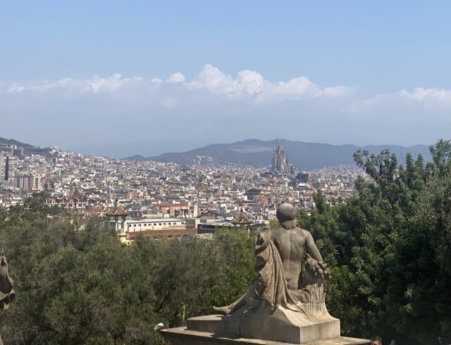 Estatua de piedra de figura recostada con drapeado pesado en primer plano, mirando hacia la ciudad. Al fondo, un mar de tejados y, asomando entre las colinas, la silueta inconfundible de una basílica inacabada con sus torres puntiagudas. La piedra moderna mira a la piedra contemporánea, ambas convencidas de su monumentalidad.