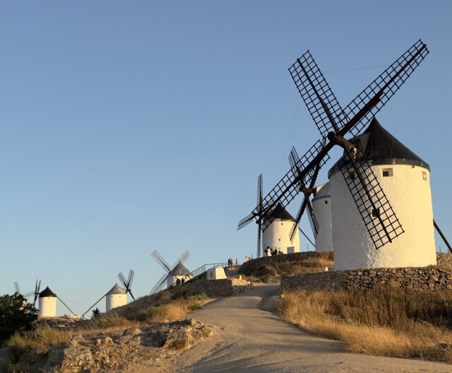 Cuatro molinos de viento blancos alineados sobre la cresta de un cerro, aspas de madera oscura recortadas contra cielo limpio del atardecer. Pequeñas figuras dispersas dan escala. Lo que un hidalgo confundió con gigantes lleva siglos siendo más bien gigantes en serio: trituraron grano hasta que la electricidad los mandó al pintoresquismo.