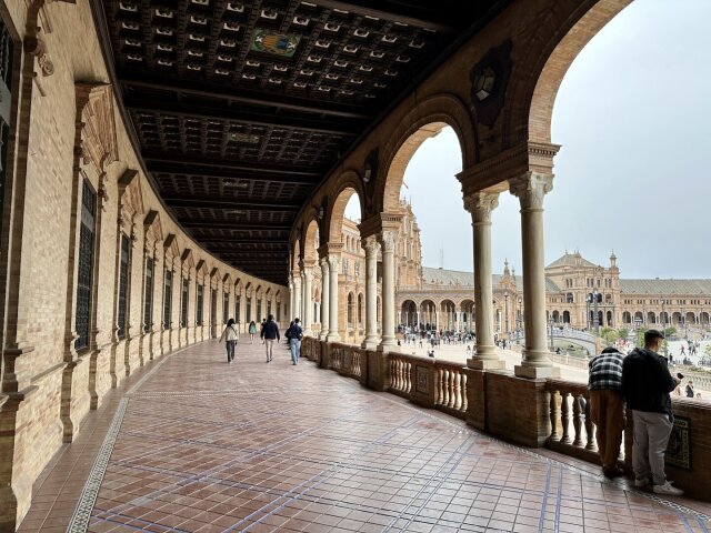 Galería interior abovedada con techo artesonado polícromo y arcos de medio punto sobre columnas pareadas, suelo de baldosa rosa. Al fondo, gente paseando bajo la curva infinita; a la derecha, la fachada de la plaza con sus torres y arquerías. Arquitectura ceremonial pensada para impresionar a los suyos y a los de fuera: a un siglo de distancia, la cámara sigue cayendo en la trampa.