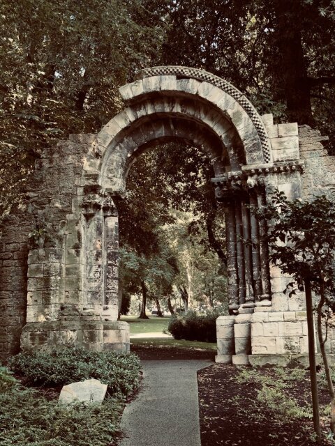 Arco románico exento en mitad de un parque, ajedrezado en la rosca, capiteles labrados sostenidos por columnas adosadas. El sendero pasa limpio por debajo. Una puerta superviviente sin muro al que pertenecer, ofreciendo umbral a quien acepte cruzar nada.