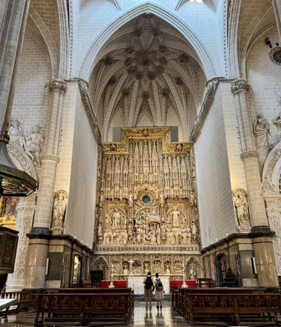 Bóvedas estrelladas sobre pilares cilíndricos y un retablo mayor que arde en oro al fondo, dos personas detenidas a su pie devueltas a su escala. Capillas laterales se asoman entre las columnas. La piedra blanca aspira hacia arriba, el oro tira hacia el mito.